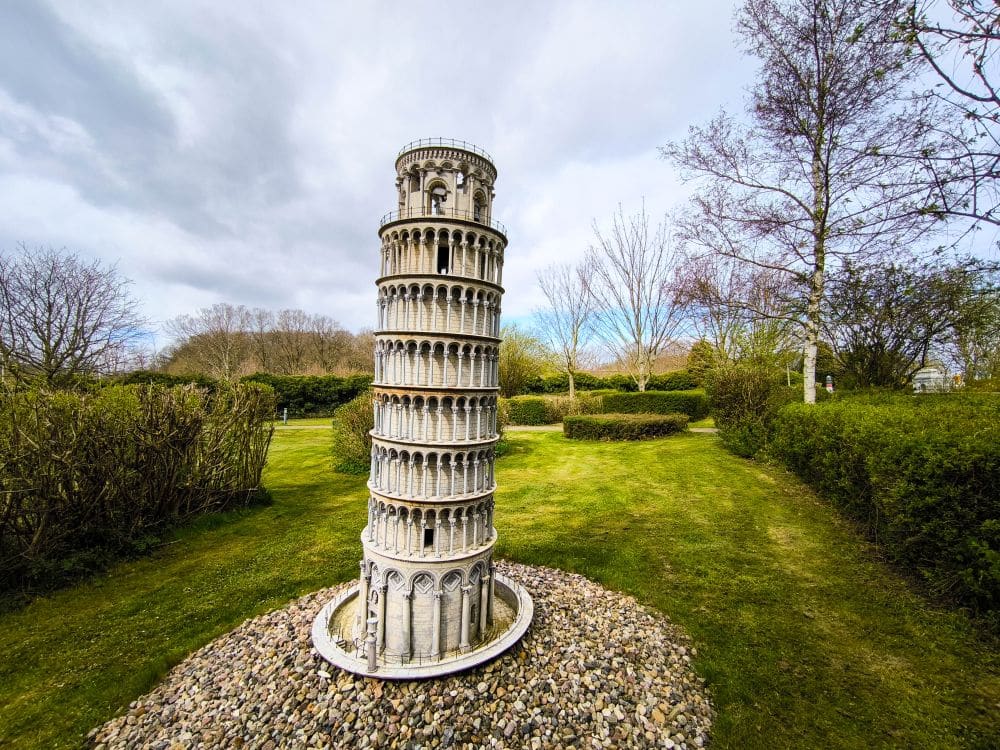 Ein kleines Modell des Schiefen Turms von Pisa steht auf einem kreisrunden Felsbett auf der Wiese im Rügenpark, umgeben von Bäumen und Sträuchern unter einem bewölkten Himmel.