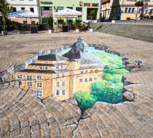 Eine realistische 3D-Straßenmalerei auf Kopfsteinpflaster in Sondershausen zeigt ein Gebäude, das sich unter dem Boden verbirgt, als wäre das Pflaster aufgebrochen.