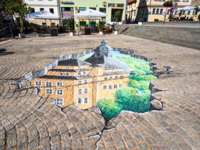 Eine realistische 3D-Straßenmalerei auf Kopfsteinpflaster in Sondershausen zeigt ein Gebäude, das sich unter dem Boden verbirgt, als wäre das Pflaster aufgebrochen.