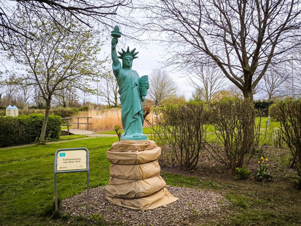 Eine kleine Nachbildung der Freiheitsstatue steht auf einem gepolsterten Sockel im Rügenpark, eingebettet in eine von Bäumen umgebene Grünanlage. Ein Schild vor der Statue informiert über dieses Wahrzeichen.