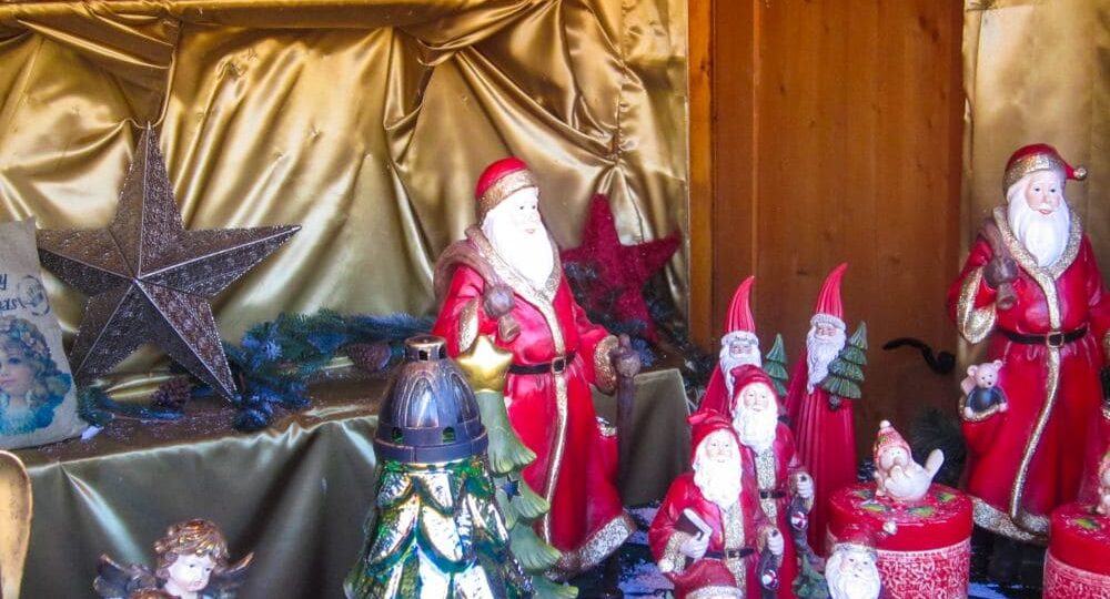 Eine festliche, von Aiderbichl inspirierte Auslage zeigt mehrere Weihnachtsmannfiguren, rote und goldene Dekorationen, silberne Sterne, Wichtel, einen Weihnachtsbaum und Engelsfiguren, die alle auf einem goldenen Stoff mit einem gemütlichen Holzhintergrund angeordnet sind.