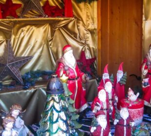 Eine festliche, von Aiderbichl inspirierte Auslage zeigt mehrere Weihnachtsmannfiguren, rote und goldene Dekorationen, silberne Sterne, Wichtel, einen Weihnachtsbaum und Engelsfiguren, die alle auf einem goldenen Stoff mit einem gemütlichen Holzhintergrund angeordnet sind.