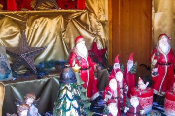 Eine festliche, von Aiderbichl inspirierte Auslage zeigt mehrere Weihnachtsmannfiguren, rote und goldene Dekorationen, silberne Sterne, Wichtel, einen Weihnachtsbaum und Engelsfiguren, die alle auf einem goldenen Stoff mit einem gemütlichen Holzhintergrund angeordnet sind.