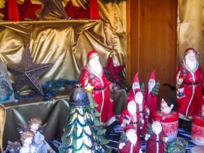 Eine festliche, von Aiderbichl inspirierte Auslage zeigt mehrere Weihnachtsmannfiguren, rote und goldene Dekorationen, silberne Sterne, Wichtel, einen Weihnachtsbaum und Engelsfiguren, die alle auf einem goldenen Stoff mit einem gemütlichen Holzhintergrund angeordnet sind.