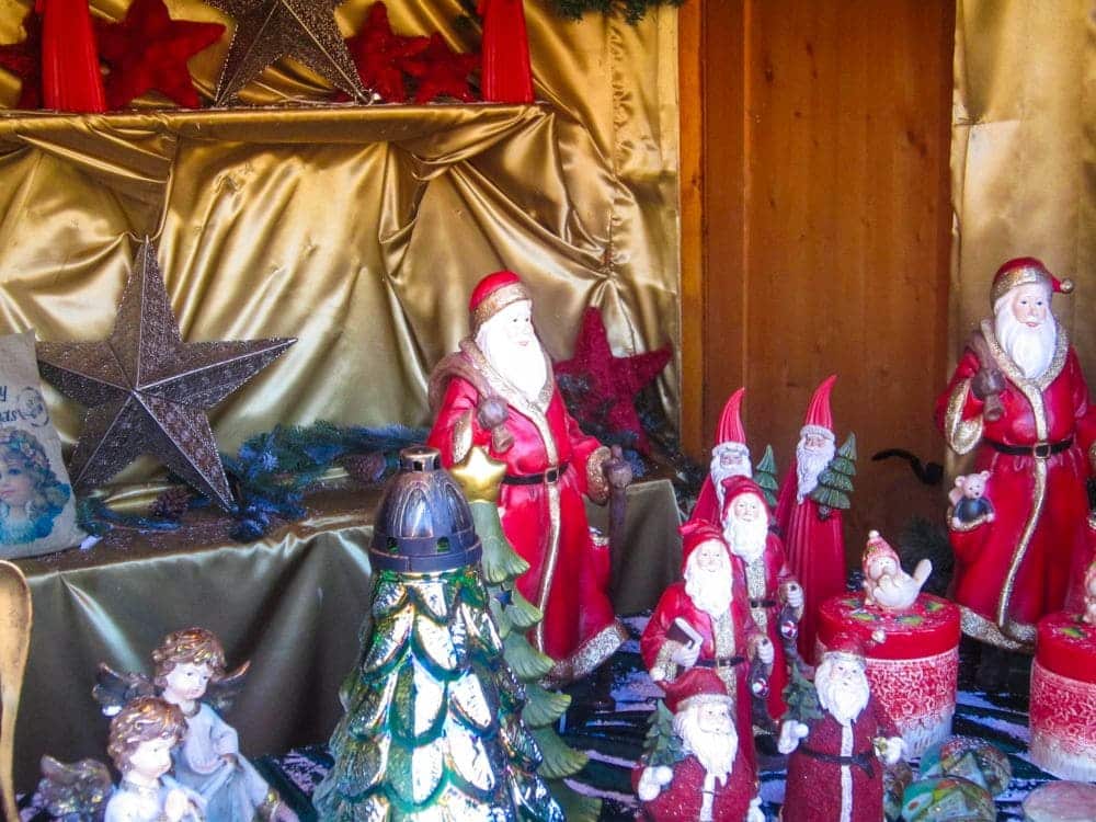 Eine festliche, von Aiderbichl inspirierte Auslage zeigt mehrere Weihnachtsmannfiguren, rote und goldene Dekorationen, silberne Sterne, Wichtel, einen Weihnachtsbaum und Engelsfiguren, die alle auf einem goldenen Stoff mit einem gemütlichen Holzhintergrund angeordnet sind.