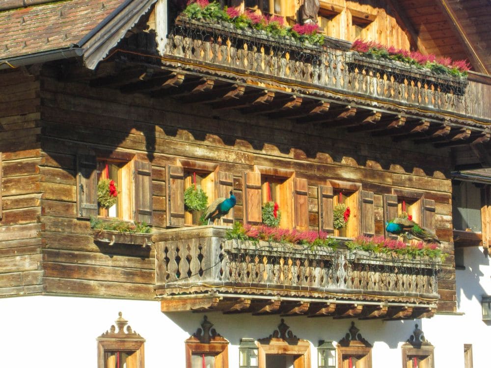 Ein rustikales Holzhaus mit blumengeschmückten Balkonen und Fensterkästen, wobei das helle Sonnenlicht die detaillierten Holzarbeiten hervorhebt. Zwei Pfauen stehen auf einem der Aiderbichl-Balkone inmitten der Blumen.