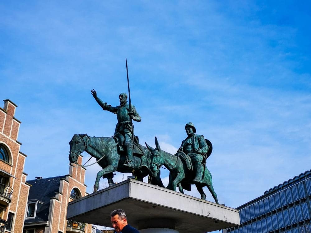 Bronzestatue von zwei auf Eseln reitenden Figuren, von denen eine ein Schwert hält und einen Arm hebt, auf einem Sockel auf einem städtischen Platz in Brüssel, mit Gebäuden und blauem Himmel im Hintergrund.