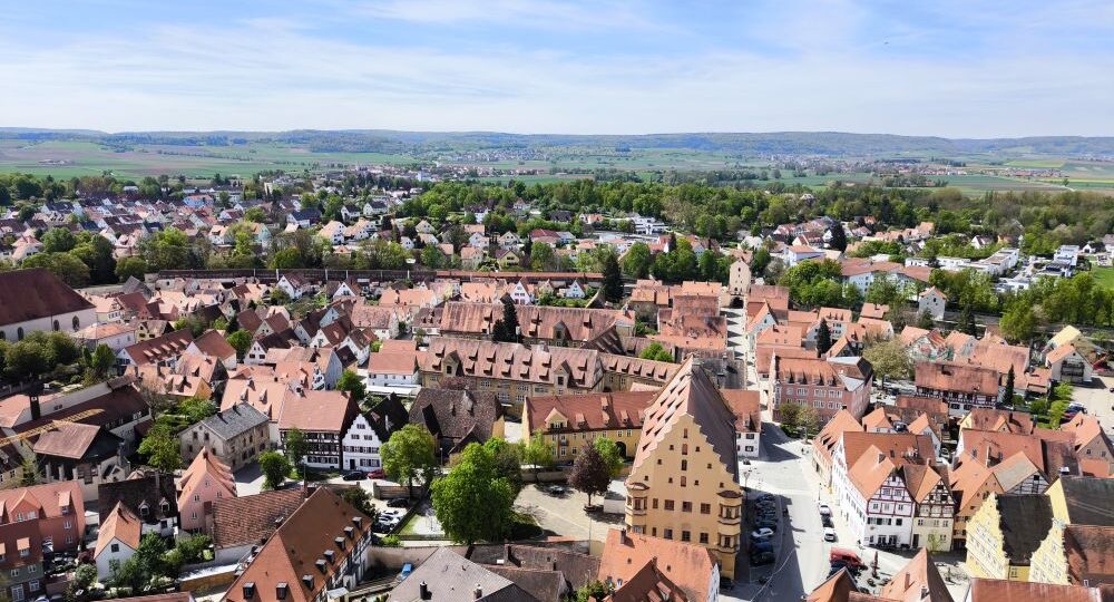 Luftaufnahme einer malerischen europäischen Stadt mit roten Ziegeldächern, engen Straßen und historischen Gebäuden, umgeben von grünen Feldern - aufgenommen von Daniel - vor entfernten Hügeln unter einem teilweise bewölkten blauen Himmel.