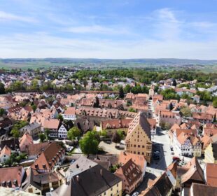 Luftaufnahme einer malerischen europäischen Stadt mit roten Ziegeldächern, engen Straßen und historischen Gebäuden, umgeben von grünen Feldern - aufgenommen von Daniel - vor entfernten Hügeln unter einem teilweise bewölkten blauen Himmel.