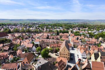 Luftaufnahme einer malerischen europäischen Stadt mit roten Ziegeldächern, engen Straßen und historischen Gebäuden, umgeben von grünen Feldern - aufgenommen von Daniel - vor entfernten Hügeln unter einem teilweise bewölkten blauen Himmel.