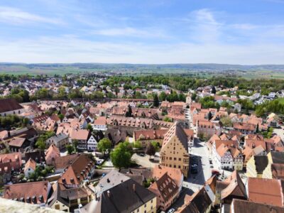 Luftaufnahme einer malerischen europäischen Stadt mit roten Ziegeldächern, engen Straßen und historischen Gebäuden, umgeben von grünen Feldern - aufgenommen von Daniel - vor entfernten Hügeln unter einem teilweise bewölkten blauen Himmel.