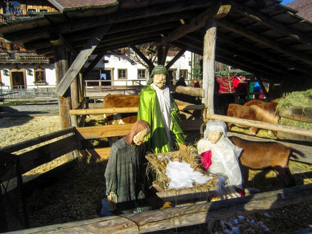 Drei Krippenfiguren - Maria, Josef und das Jesuskind in einer Krippe - stehen unter einer hölzernen Stallkonstruktion in Aiderbichl, im Hintergrund Kühe und Heu, alles hell erleuchtet vom Sonnenlicht.
