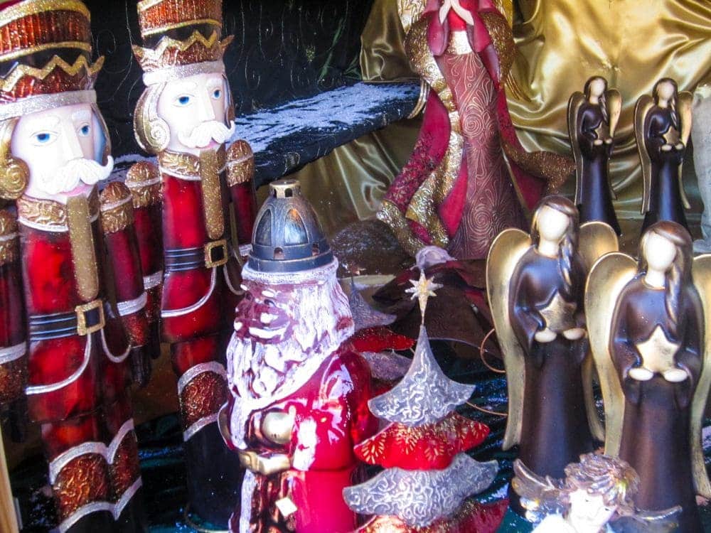 Eine festliche Auslage mit Nussknackerfiguren, einem verzierten Weihnachtsmann, goldenen Engeln, einem dekorativen Weihnachtsbaum und einer gewandeten Figur vor einem gold-blauen Hintergrund, der an Aiderbichls charmante Weihnachtsszenen erinnert.