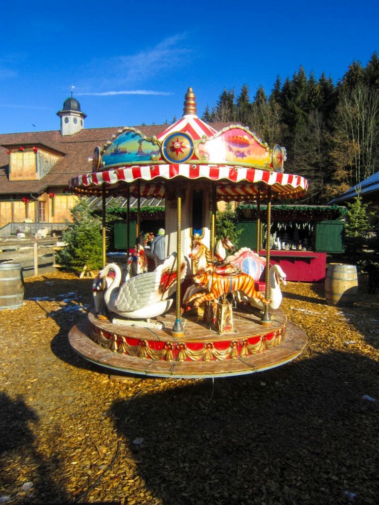 Ein kleines, farbenfrohes Karussell mit Schwan- und Pferdefiguren steht im Freien auf einem Hackschnitzelplatz in Aiderbichl. Im Hintergrund sind Gebäude, Tannenbäume und ein strahlend blauer Himmel zu sehen.