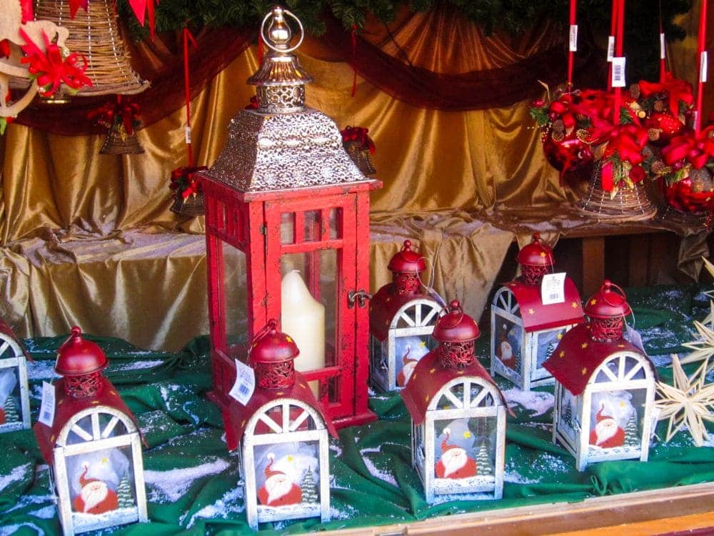 Eine festliche Auslage aus roten Laternen, jede mit einem Weihnachtsmannmotiv, umgibt eine größere Laterne mit einer weißen Kerze auf der Aiderbichl-Weihnachtsveranstaltung. Die Dekoration besteht aus goldenem und grünem Stoff, weihnachtlichen Bändern und kleinen dekorativen Akzenten.