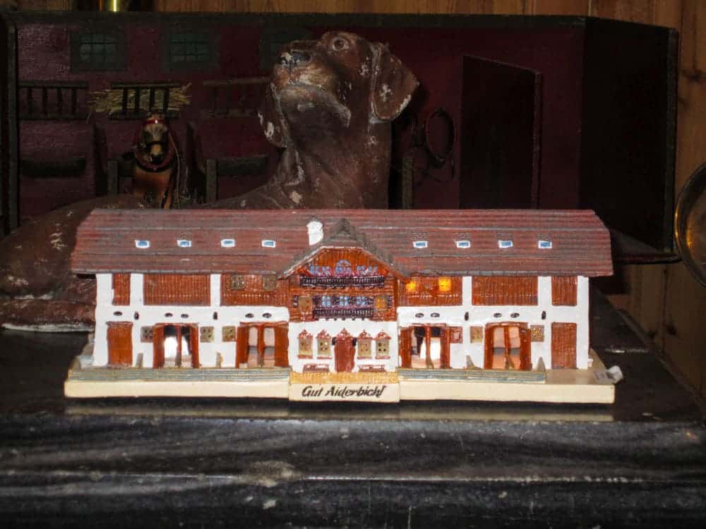 Ein Modell eines traditionellen Chalets mit der Aufschrift "Gut Aiderbichl" steht vor einer großen, realistischen Hundeskulptur, die den einladenden Geist von Aiderbichl in einem warm beleuchteten Raum einfängt.