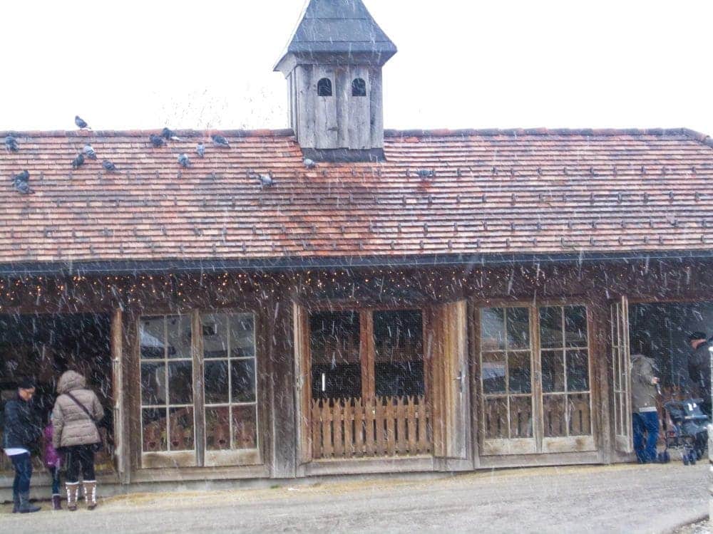 Ein hölzernes Aiderbichl-Gebäude mit Ziegeldach und Kuppel, geschmückt mit Lichterketten. Es schneit, Menschen stehen unter dem Vordach und Tauben sitzen auf dem Dach.