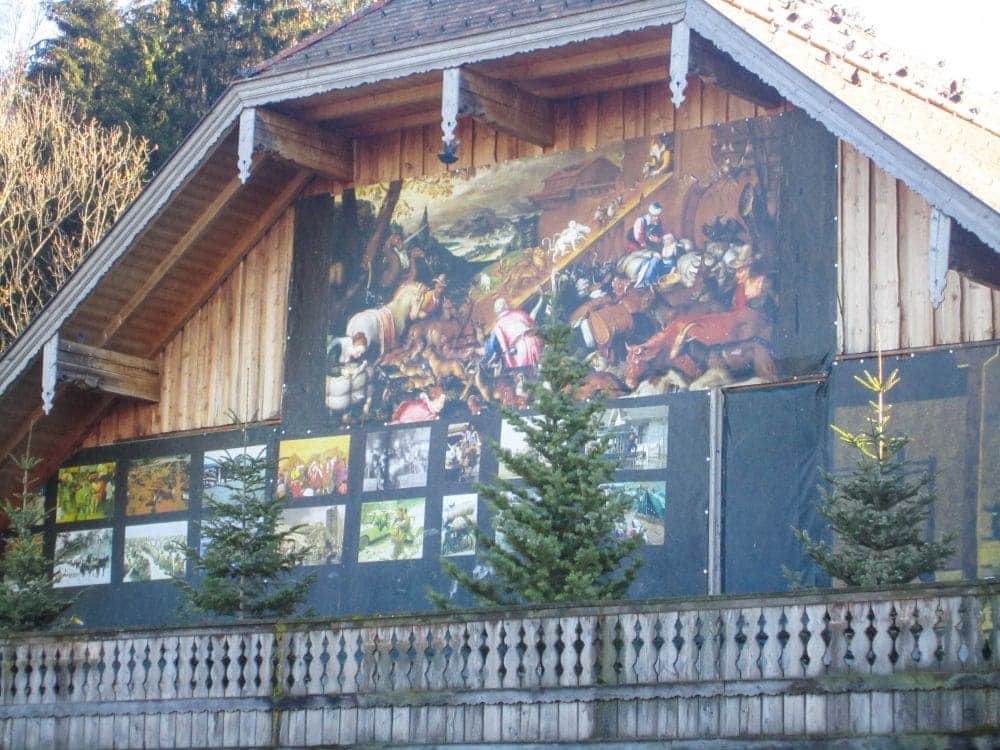 An der Holzfassade eines Gebäudes in Aiderbichl befindet sich ein großes Wandbild, das eine dramatische historische Schlachtszene darstellt. Darunter sind mehrere kleinere Bilder angeordnet, davor stehen immergrüne Bäume.