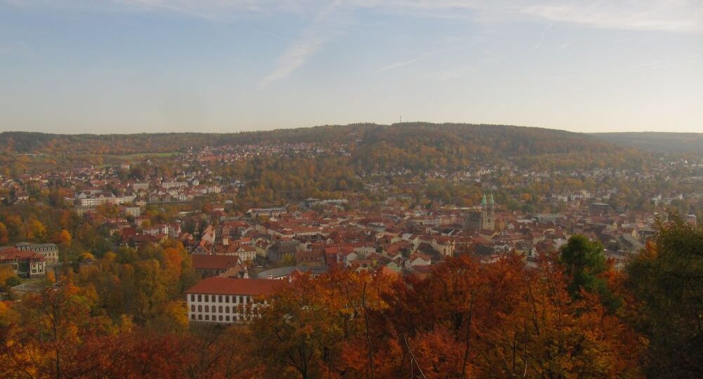 In der Ferne bietet sich ein Panoramablick auf den Dreißigacker mit seinen roten Dächern, umgeben von herbstlich gefärbten Bäumen - perfekt für eine Herbstwanderung unter dem teilweise bewölkten Himmel, bei der das Dietzhäuschen inmitten sanfter Hügel zu sehen ist.