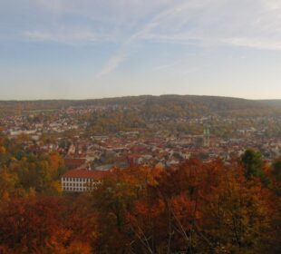 In der Ferne bietet sich ein Panoramablick auf den Dreißigacker mit seinen roten Dächern, umgeben von herbstlich gefärbten Bäumen - perfekt für eine Herbstwanderung unter dem teilweise bewölkten Himmel, bei der das Dietzhäuschen inmitten sanfter Hügel zu sehen ist.