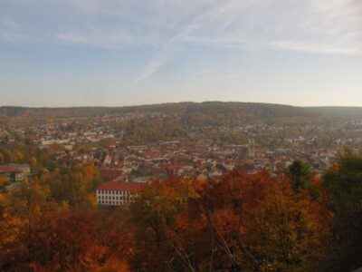 In der Ferne bietet sich ein Panoramablick auf den Dreißigacker mit seinen roten Dächern, umgeben von herbstlich gefärbten Bäumen - perfekt für eine Herbstwanderung unter dem teilweise bewölkten Himmel, bei der das Dietzhäuschen inmitten sanfter Hügel zu sehen ist.