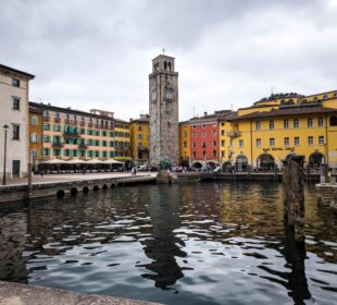 Bunte Gebäude und der hohe steinerne Uhrenturm von Riva del Garda säumen die Uferpromenade, deren Spiegelungen im Kanal schimmern, während die Menschen bei bewölktem Himmel an der Promenade flanieren.