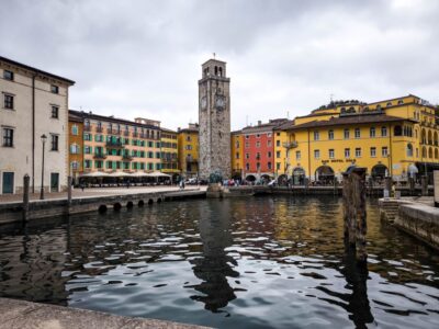Bunte Gebäude und der hohe steinerne Uhrenturm von Riva del Garda säumen die Uferpromenade, deren Spiegelungen im Kanal schimmern, während die Menschen bei bewölktem Himmel an der Promenade flanieren.