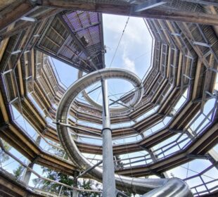 Blick nach oben durch den Abenteuer-Spiralholzsteg des Baumwipfelpfads Bad Wildbad, mit Metallgeländer und einer zentralen, sich windenden Silberrutsche, offen zum blauen Himmel mit Wolken darüber.