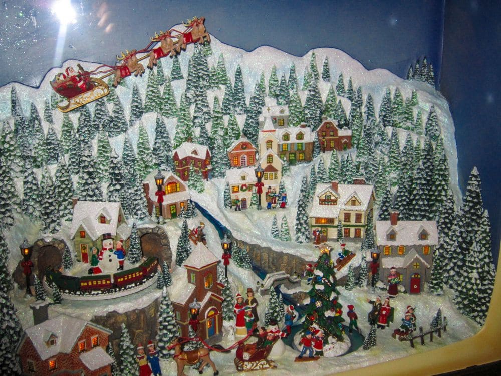 Eine detaillierte Miniatur-Weihnachtsdorfszene mit schneebedeckten Häusern, Tannenbäumen, feiernden Menschen und dem Schlitten des Weihnachtsmanns mit Rentieren, die darüber hinwegfliegen - das erinnert an den festlichen Charme von Aiderbichl im Winter.