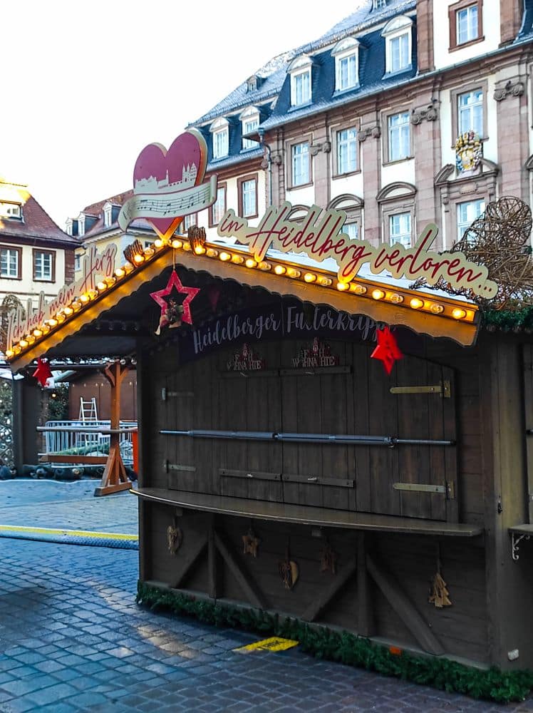 Ein hölzerner Weihnachtsmarktstand, geschmückt mit Lichtern, roten Sternen und festlichen Girlanden, steht geschlossen auf einer Kopfsteinpflasterstraße vor historischen Gebäuden in Heidelberg, Deutschland.