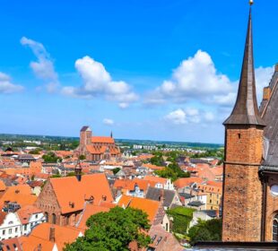 Luftaufnahme von Wismar, einer europäischen Stadt mit roten Dächern, einer großen Kirche in der Ferne und einem hohen Backsteinturm auf der rechten Seite unter einem strahlend blauen Himmel mit vereinzelten Wolken.