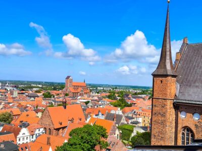 Luftaufnahme von Wismar, einer europäischen Stadt mit roten Dächern, einer großen Kirche in der Ferne und einem hohen Backsteinturm auf der rechten Seite unter einem strahlend blauen Himmel mit vereinzelten Wolken.