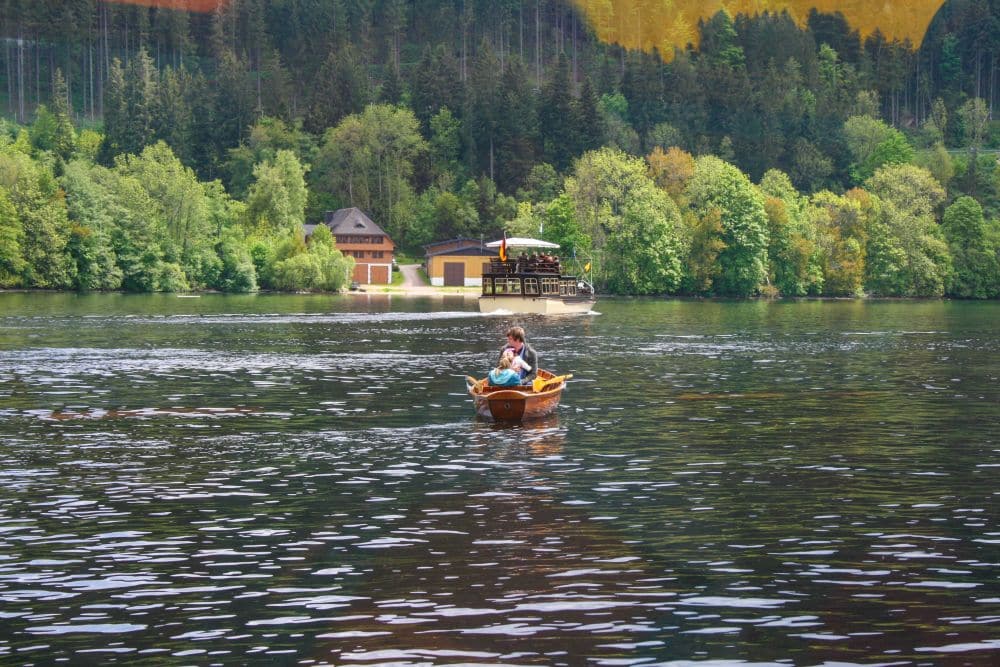 Ein kleines hölzernes Ruderboot mit zwei Personen schwimmt auf dem ruhigen Wasser des Titisees, umgeben von dichten grünen Bäumen. Im Hintergrund stehen Gebäude in Ufernähe und ein größeres Boot gleitet über den See.