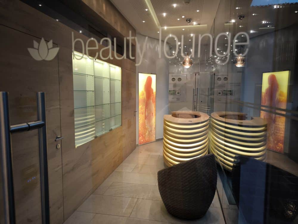 Eine moderne Kastel-Beauty-Lounge mit Glastüren, beleuchteter Beschilderung, stilvollen spiralförmigen Empfangsschaltern, gedämpfter Beleuchtung und künstlerischen Wandpaneelen mit abstrakten orangefarbenen Motiven.