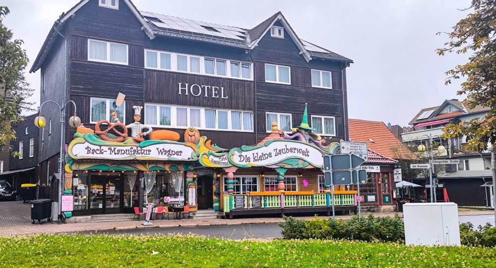Hinter einer bunten Bäckerei und einem Süßwarenladen in Braunlage liegt an einem bewölkten Tag ein dreistöckiges Hotel aus dunklem Holz mit weißer Verkleidung, davor Grünflächen und eine kleine Straße.