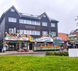 Hinter einer bunten Bäckerei und einem Süßwarenladen in Braunlage liegt an einem bewölkten Tag ein dreistöckiges Hotel aus dunklem Holz mit weißer Verkleidung, davor Grünflächen und eine kleine Straße.