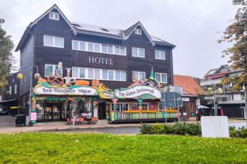 Hinter einer bunten Bäckerei und einem Süßwarenladen in Braunlage liegt an einem bewölkten Tag ein dreistöckiges Hotel aus dunklem Holz mit weißer Verkleidung, davor Grünflächen und eine kleine Straße.