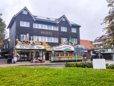 Hinter einer bunten Bäckerei und einem Süßwarenladen in Braunlage liegt an einem bewölkten Tag ein dreistöckiges Hotel aus dunklem Holz mit weißer Verkleidung, davor Grünflächen und eine kleine Straße.