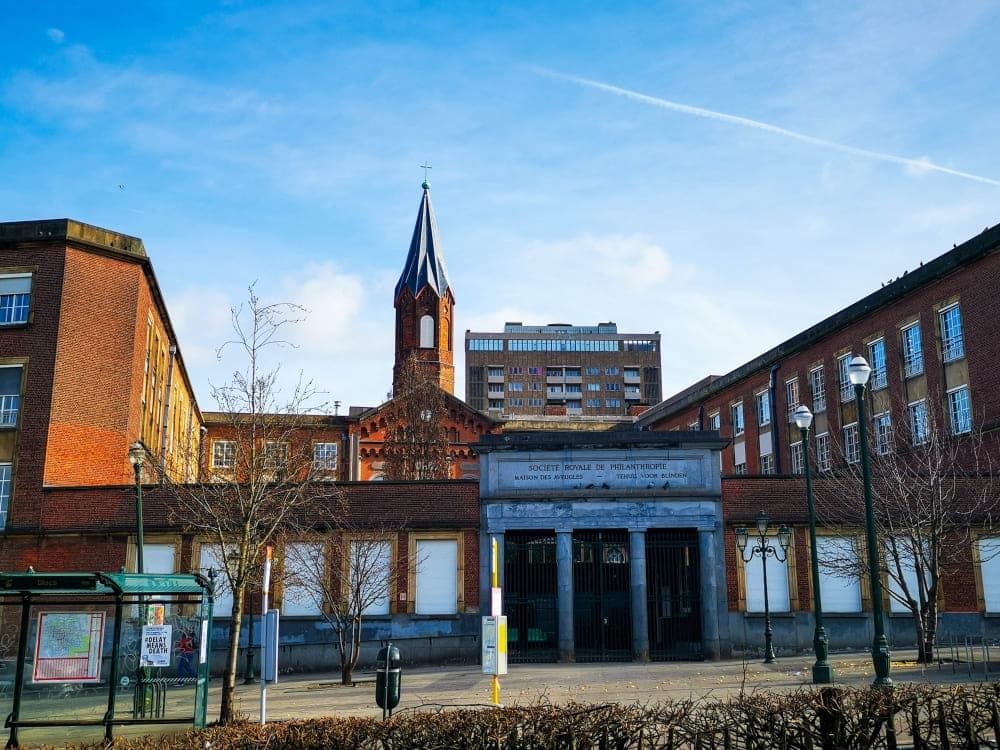 Ein historisches rotes Backsteingebäude mit zentralem Turm und Bogenfenstern steht hinter einer Toreinfahrt in Brüssel; im Hintergrund sind kahle Bäume und ein modernes Wohnhaus unter blauem Himmel zu sehen.