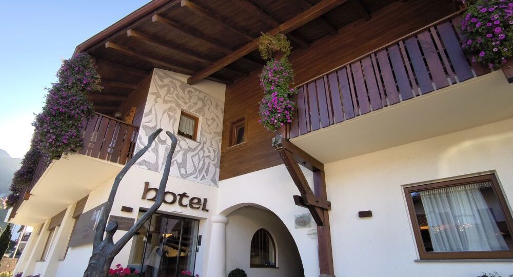 Ein Hoteleingang mit Holzbalkonen, hängenden Blumenkörben und großen Fenstern. Das Wort Kastel steht über der Glastür. Das Gebäude verfügt über ein Wandgemälde und Topfpflanzen am Eingang.