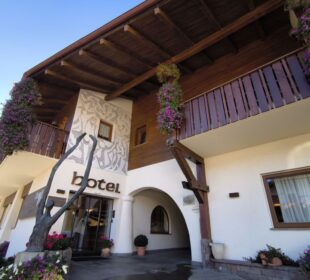 Ein Hoteleingang mit Holzbalkonen, hängenden Blumenkörben und großen Fenstern. Das Wort Kastel steht über der Glastür. Das Gebäude verfügt über ein Wandgemälde und Topfpflanzen am Eingang.