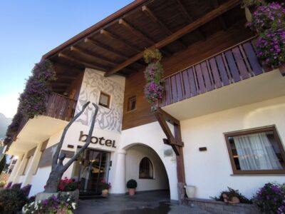 Ein Hoteleingang mit Holzbalkonen, hängenden Blumenkörben und großen Fenstern. Das Wort Kastel steht über der Glastür. Das Gebäude verfügt über ein Wandgemälde und Topfpflanzen am Eingang.