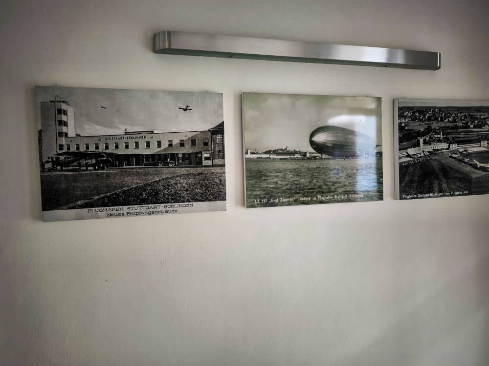 Drei Schwarz-Weiß-Fotografien aus der Luftfahrt sind auf einer weißen Wand unter einer modernen Metallleuchte ausgestellt. Die Bilder zeigen den Flughafen Böblingen, ein Luftschiff und historische Flugplätze.