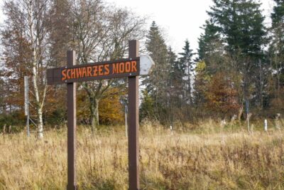 Ein hölzernes Schild mit der Aufschrift "Schwarzes Moor" steht auf einer grasbewachsenen Wiese, umgeben von herbstlich gefärbten Bäumen und immergrünen Bäumen unter einem leicht bedeckten Himmel und markiert den Eingang zu diesem malerischen Moorgebiet.