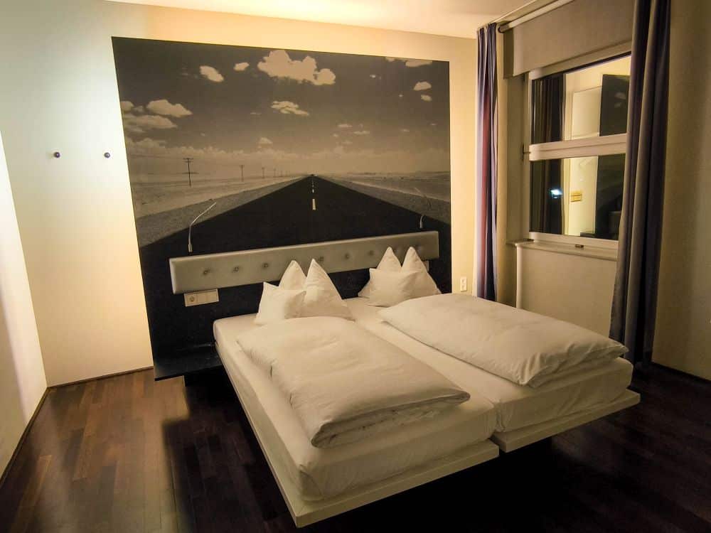 Ein modernes Hotelzimmer in Böblingen verfügt über ein Doppelbett mit weißem Bettzeug, Holzböden, neutrale Wände und ein großes Wandgemälde einer leeren Straße, die sich bis zum Horizont erstreckt. Natürliches Licht erfüllt den Raum durch ein Fenster mit Vorhängen.