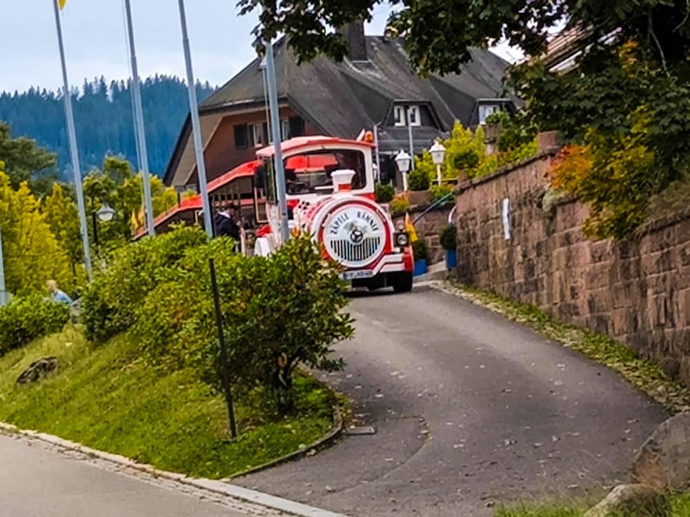 Ein kleiner rot-weißer Touristenzug fährt eine schmale Straße in Titisee hinauf, vorbei an üppigem Grün und Häusern, mit einer Steinmauer auf der einen Seite und Fahnen auf der linken Seite.