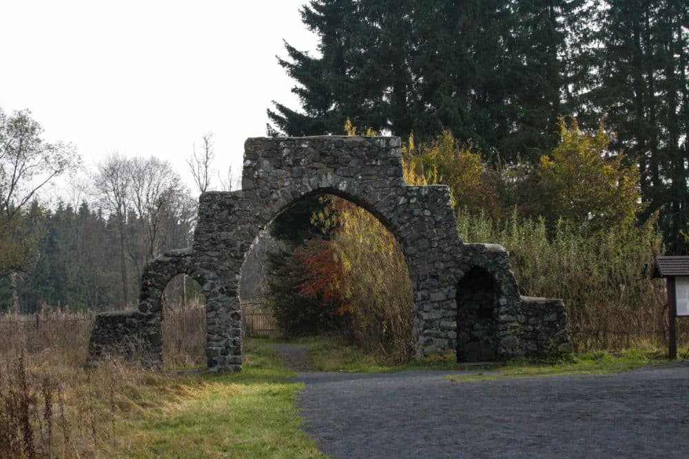 Ein steinerner Torbogen mit drei ungleichmäßigen Bögen steht auf einem Kiesweg, der von Gras, Sträuchern und hohen Bäumen umgeben ist und den zeitlosen Charme eines Moors in einer ruhigen, natürlichen Umgebung hervorruft.