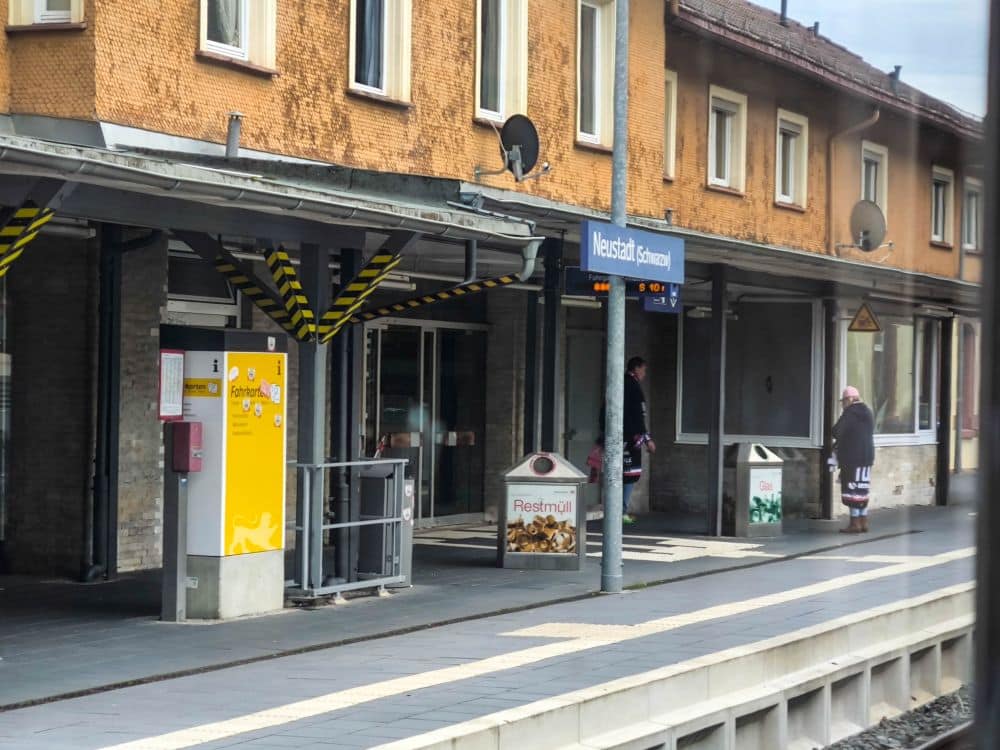 Ein Bahnsteig mit einem gelben Verkaufsautomaten, Mülleimern und Menschen, die in der Nähe des Gebäudes stehen. Auf dem Bahnhofsschild steht "Wesel-Stadt" - eine Anspielung auf Kleinstädte wie Titisee - und der Bahnsteig ist bis auf ein paar Personen weitgehend leer.