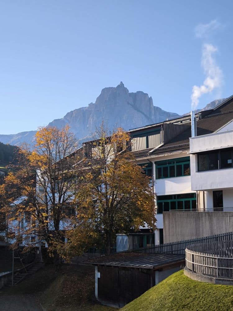 Weiße, moderne Gebäude mit grünen Fenstern und herbstlichen Bäumen stehen im Vordergrund von Kastel, während sich im Hintergrund ein hoher, felsiger Berg unter einem klaren blauen Himmel erhebt.