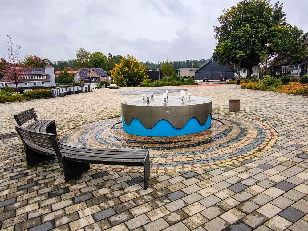 Ein runder Brunnen mit blauen Wellenmustern und kleinen Wasserdüsen steht an einem bewölkten Tag auf einem gepflasterten Platz mit Bänken und Bäumen in Braunlage; im Hintergrund sind Gebäude und Grünanlagen zu sehen.